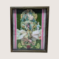 A vertical shadow box frame containing a Victorian-style paper collage. Features a cherub, floral lithographs, and a large pink velvet bow against a forest green velvet background. Housed in a dark wood-grain frame.