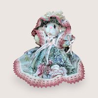 A seated plush lamb in a wide, floral-patterned sage green and rose pink dress. The lamb features long eyelashes, a pink nose, and an oversized matching lace bonnet. The intricate pink lace trim on the hem and bonnet adds a grounded, romantic texture.