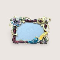 A horizontal photo frame with a deep red enamel border. Ornate gold-tone decorations include a crystal-studded purple hibiscus-style flower on the top left and a fashionable stiletto shoe on the bottom right.