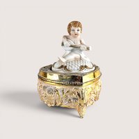 A vintage footed trinket box with a highly detailed gold-toned floral base and a white bisque-style lid featuring a cherub playing a stringed instrument.