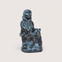 Dark carved stone sculpture of a figure in a parka standing beside an animal, centered on a neutral background.