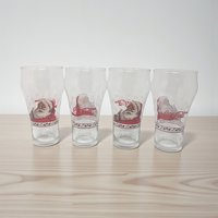 A horizontal view of four clear glass tumblers featuring the iconic Haddon Sundblom Santa Claus holding a glass of Coca-Cola. The design includes a festive holly and berry border around the base and white snowflake accents throughout the glass.