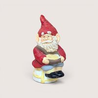 A vintage ceramic gnome figurine with a tall red hat, white beard, and glasses, sitting on a stool and holding a golden bowl.