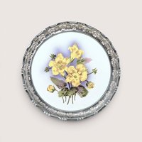 A round porcelain plaque with painted yellow flowers and a pierced silver rim, centered on a beige background.