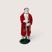 An elderly man doll wearing a red robe, spectacles, and a nightcap on a stand, centered on a beige background.