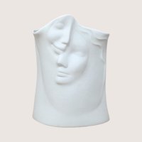 A matte white ceramic vase molded with two abstract interlocking faces, centered on a beige background.