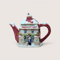 A ceramic teapot shaped like a snow-covered holiday house with festive garland, centered on a beige background.