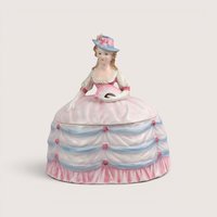 A vintage porcelain crinoline lady powder box figurine holding a fan, centered on a beige background.