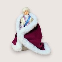 A vintage royal doll wearing a fur-trimmed purple robe and white hat, centered on a beige background.