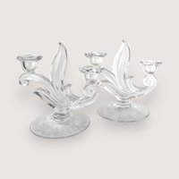 A pair of elegant clear glass candelabras with curved leaf details and flower-shaped cups, centered on a beige background.