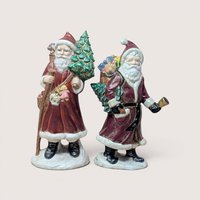 Pair of vintage Santa Claus figurines carrying Christmas trees and sacks of toys, centered on a plain linen background.