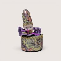 Vintage porcelain jester doll emerging from a matching round metallic fabric box with gold trim, centered on a plain linen background.
