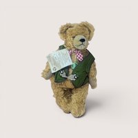 Vintage mohair teddy bear wearing a traditional Bavarian green vest and red-and-white checkered bow tie, complete with its original hanging paper tag, centered on a plain linen background.