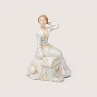 Vintage porcelain lady figurine sitting atop a small trunk or chest patterned with gold trim, featuring a white starburst-accented dress with a matching wide-brimmed hat resting at her side, centered on a plain linen background.