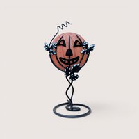 Vintage frosted orange glass jack-o'-lantern face insert sitting inside a black metal wire frame stand accented with leaves and a curly vine top, centered on a plain linen background.