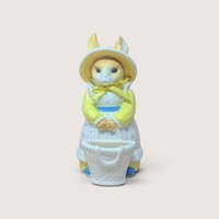 Vintage ceramic bunny figurine dressed in a yellow long-sleeved shirt, a yellow bonnet with a bow, and a blue polka-dot apron dress, standing behind a small woven white basket, centered on a plain linen background.