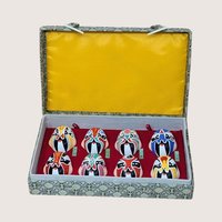 A vintage boxed display of eight hand painted Beijing Opera clay masks resting on a soft pink background. The vibrant, colorful theatrical facial makeup designs are arranged on red velvet inside a fabric covered case with a bright yellow satin lined lid.