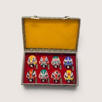 Boxed set of eight vintage miniature Chinese opera masks, hand-painted with vibrant traditional designs and arranged in two rows of four inside a red-lined display case with a bright yellow fabric lid interior, centered on a plain linen background.