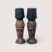 Pair of vintage hand-carved wooden candlesticks depicting stylized elongated faces with carved hair details, each supporting a dark-stained cup-style candle holder accented with geometric chevron patterns, centered on a plain linen background.