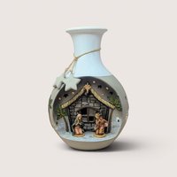 Vintage ceramic vase featuring a built-in, recessed Nativity scene on the front, depicting Mary and Joseph kneeling inside a stone stable flanked by palm trees under cutout star accents, complete with a white glazed upper neck wrapped in natural twine with hanging clay stars, centered on a plain linen background.