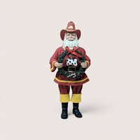 Vintage standing Santa Claus figurine dressed as a firefighter in a red turnout coat with yellow trim, black boots, and a matching red "FD" firefighter hat, holding a green Christmas wreath adorned with a red bow, a candy cane, and a black-and-white spotted dog bone ornament, centered on a plain linen background.