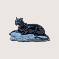 A vintage hand painted ceramic black panther figurine on a soft pink background. Made in 1994, the glossy black big cat sits in a reclined, alert pose atop a matte, textured grey rock base.