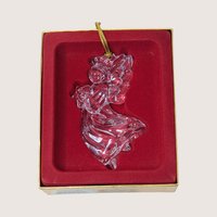 A vintage Gorham crystal angel ornament with a gold tassel on a soft pink background. The fine German glass sculpture features a clear, faceted angel in flight holding a small star, catching the light with brilliant clarity.