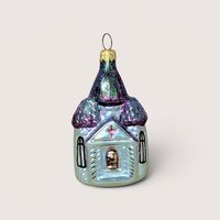 A vintage Christopher Radko blown glass church ornament with its box on a soft pink background. The fine mercury glass cathedral features iridescent pale green walls, hand painted black windows, and shimmering purple steeples covered in sparkling glitter.