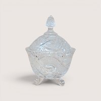 A vintage clear crystal candy dish with a matching lid and three scroll feet on a plain background