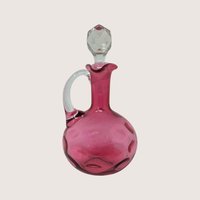 Cranberry glass cruet bottle with a clear applied handle and a faceted clear glass stopper against a neutral background