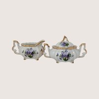 A vintage porcelain creamer and lidded sugar bowl set featuring delicate purple violet bouquets and gold trim.