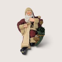 Vintage holiday figurine of Santa Claus sitting down, checking a long list with a pen and holding a toy bag.