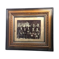 A large, ornate antique wood and gesso picture frame with a gold-toned finish and deep molding, housing a sepia-toned historical photograph of people in formal Victorian attire.