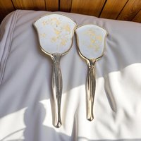 A vertical view of a vintage hand mirror and a matching hairbrush featuring an ornate silver-toned handle and frame. The backs of both pieces feature a white background decorated with a delicate gold-etched pattern of birds perched on flowering branches. The mirror has a wide, scalloped head, while the brush has a matching tapered shape.