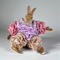 A vertical view of a vintage figural rabbit ornament with a hand-painted resin head and paws. The rabbit is dressed in an elaborate costume featuring ruffled pink and lavender satin sleeves, a white lace collar, and voluminous champagne-colored satin pants with gold-painted shoes.