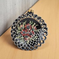 A 4-inch round black clay ornament with a delicate reticulated or pierced lattice edge, featuring hand-painted red and white flowers with green leaves.