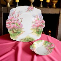 Scalloped square plate and a small dual-handled bonbon dish featuring a lush landscape of blossoming pink cherry trees. The porcelain displays a watercolor-style scene with vibrant pink florals, green grass, and a winding path against a white ground.
