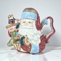 Ceramic teapot in the shape of a traditional St. Nicholas figure wearing a red hooded robe with white trim. He holds a small green Christmas tree that serves as the spout. The texture is a smooth, glossy glaze with a curved handle shaped like a candy cane.