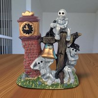 Ceramic clock tower figurine with a distressed stone texture. Features a central clock face and a white skeleton climbing the side. Vertical, rectangular silhouette with a peaked roof. Dark grey and orange accents throughout.