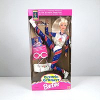 The Olympic Gymnast Barbie is pictured in her original packaging, posed in a dynamic split. She wears a patriotic red, white, and blue leotard with star accents and carries a white gym bag. A pink tumbling ring and gold medal accessory are visible within the box.