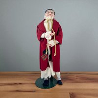 Porcelain character doll of Ebenezer Scrooge with a hand-painted, expressive face and wire-rimmed spectacles. He wears a tattered-hem white nightshirt under a long, deep red robe tied with a gold-toned rope cord. The doll features a mix of smooth glazed porcelain and soft, textured fabric.