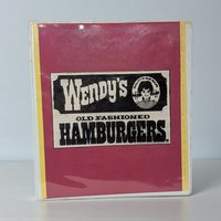 A vintage three-ring binder with a pink and white cover featuring the original Wendy's Old Fashioned Hamburgers logo. The cover includes the iconic illustration of a young girl with red pigtails and a striped shirt, set against a black and white rectangular background with yellow side borders.