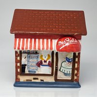 A ceramic Mrs. Fields cookie shop cookie jar. The jar is shaped like a storefront with a brown shingled roof, a red and white striped awning, and a red dome awning with the Mrs. Fields logo. The front depicts a baker inside among trays of cookies.