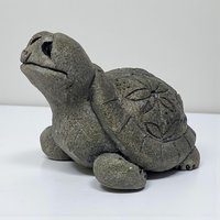 A small, heavy-textured stone turtle figurine with a weathered grey finish. The shell is embellished with a carved floral or sand dollar motif, and the figure is depicted with an upturned head and simple, rounded limbs.
