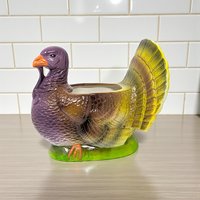 Handpainted ceramic turkey planter featuring a purple head, orange beak, and variegated brown and yellow feathers. The bird sits on a bright green base with a glossy glazed finish and a hollow center.