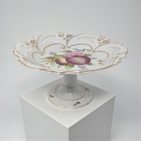A white porcelain pedestal dish featuring a hand-painted plum and pear motif at the center. The shallow bowl has a decorative pierced or reticulated rim accented with gold filigree and a matching footed base.