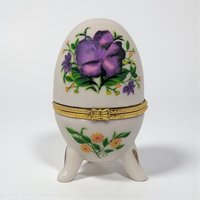 A small, egg-shaped white ceramic box with a hinged gold metal closure. The exterior is decorated with delicate purple violet sprays and green leaves on both the lid and base.
