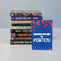 A stack of nine vintage mass-market paperbacks sits vertically against a neutral background, with a tenth book, Science Fiction by Asimov, leaning against the front. The spines show titles from authors like Roger Zelazny and Frederik Pohl. The front book features a vibrant blue grid pattern with red geometric squares.