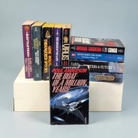 A collection of mass-market science fiction paperbacks is arranged on a white platform. A single book titled The Boat of a Million Years stands in the foreground, featuring a silver spacecraft against a dark blue and black galactic void. The surrounding books display a variety of colorful spines in shades of purple, yellow, and blue, showing visible shelf wear and creased edges along their paper bindings.