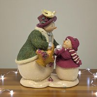 Matte resin figurine of two women in winter coats and hats walking side-by-side. One wears a green coat with a red scarf, the other a red coat with a red scarf.